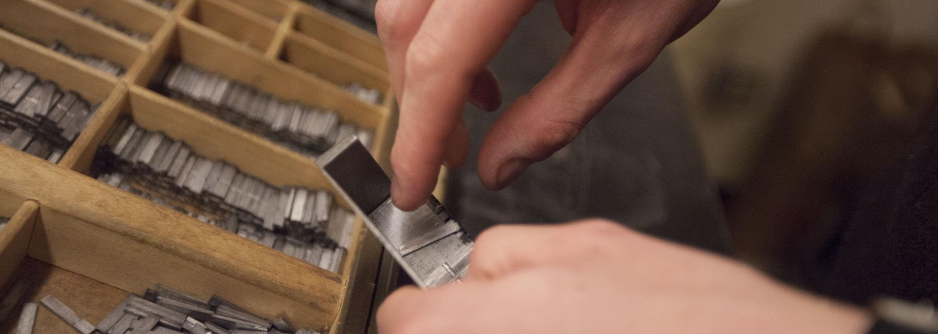 Closeup of hands arranging type pieces and compartments filled with collections of movable type.